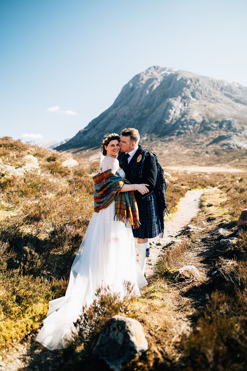 Wedding couple wrapped in tartan plaid against rugged Scottish mountain landscape.