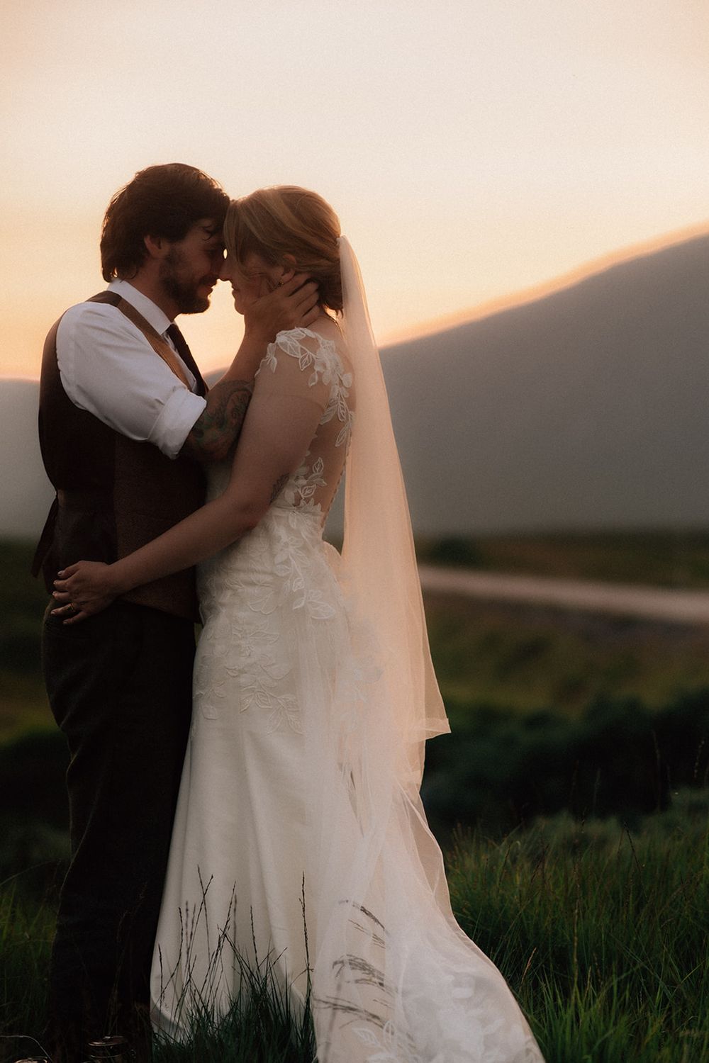 A bride and groom share an intimate moment at sunset in a lush green field with mountains in the background.