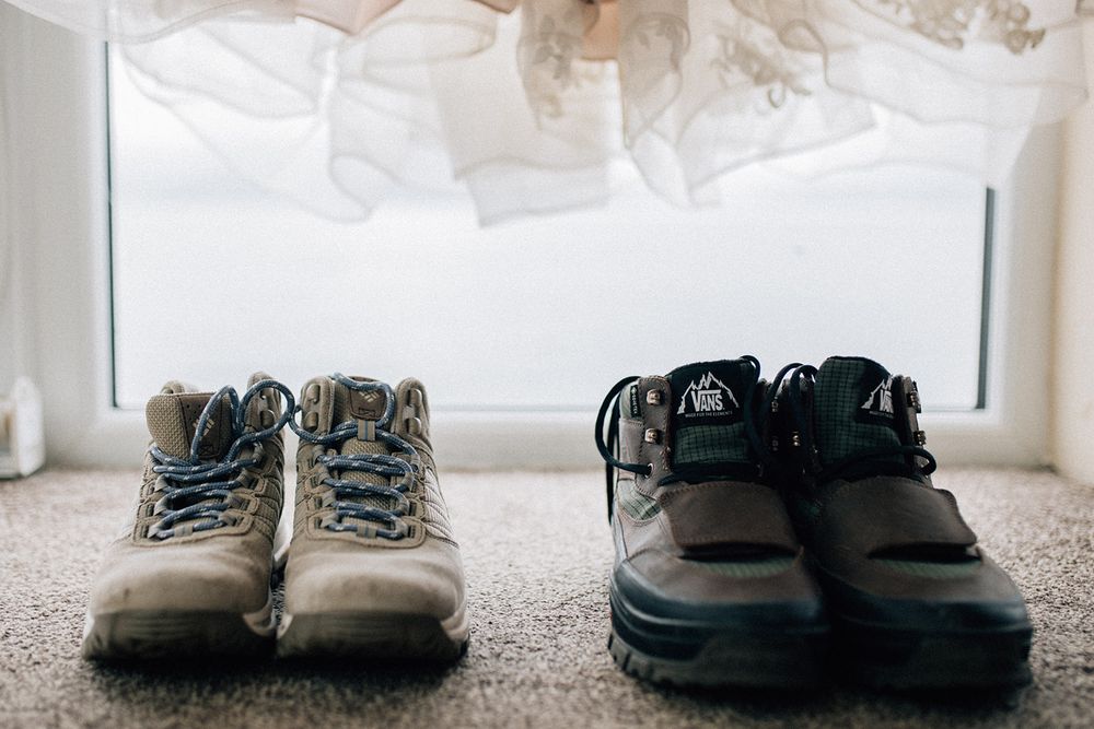 Two pairs of worn hiking boots placed side by side on carpet near a window with white fabric backdrop.