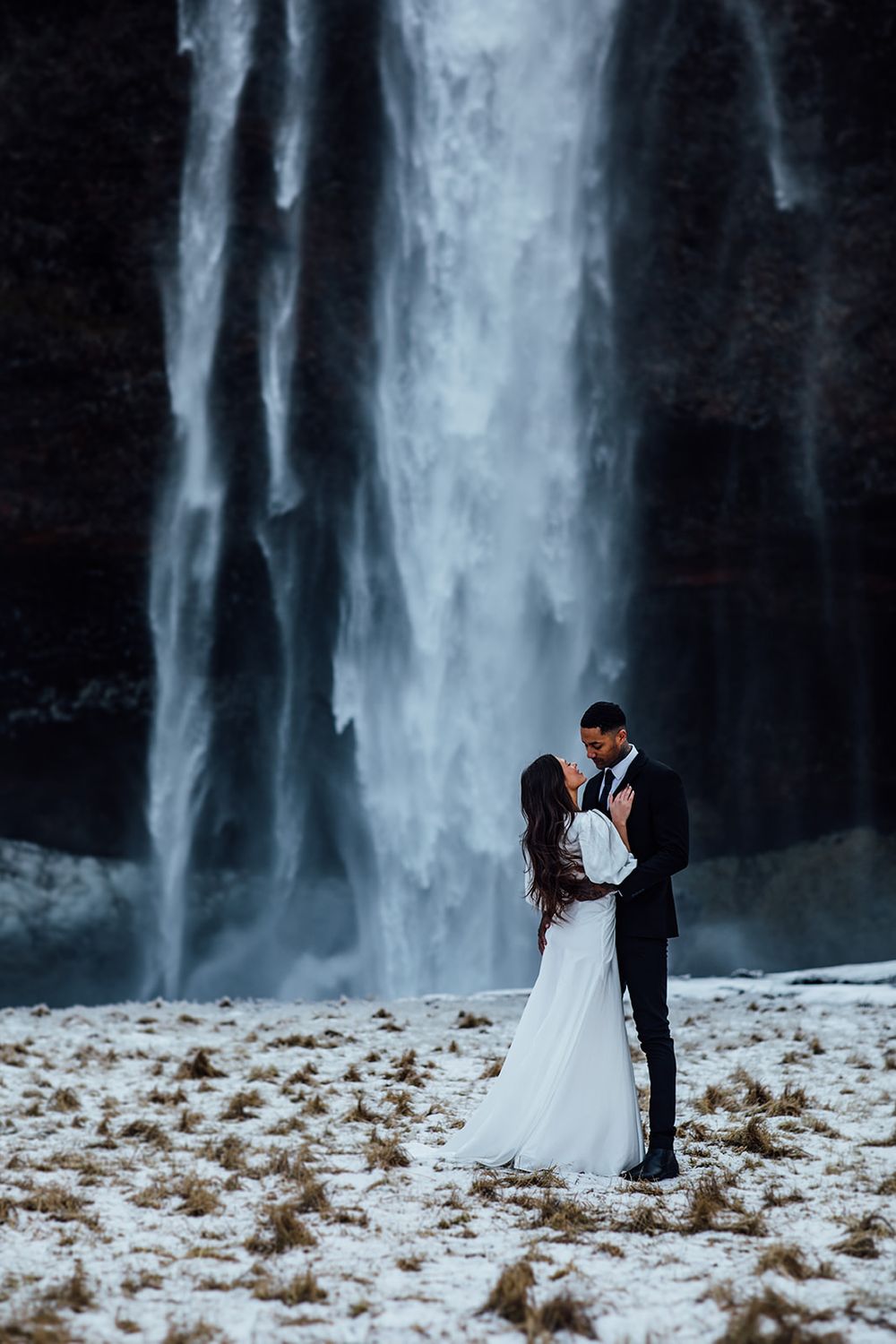 Dramatic wedding portrait in front of a towering waterfall with misty atmosphere.