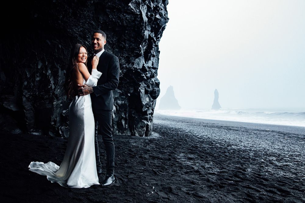 Newlyweds pose on dramatic black sand beach with rocky cliffs in the background.
