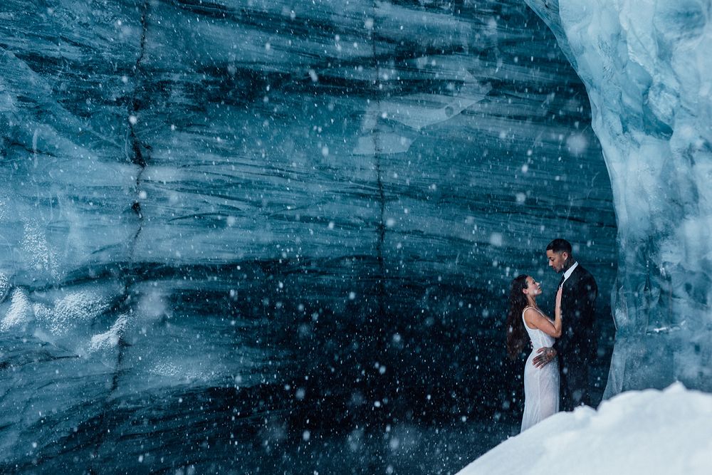 Wedding couple embraces in front of an icy blue glacier wall during snowfall.