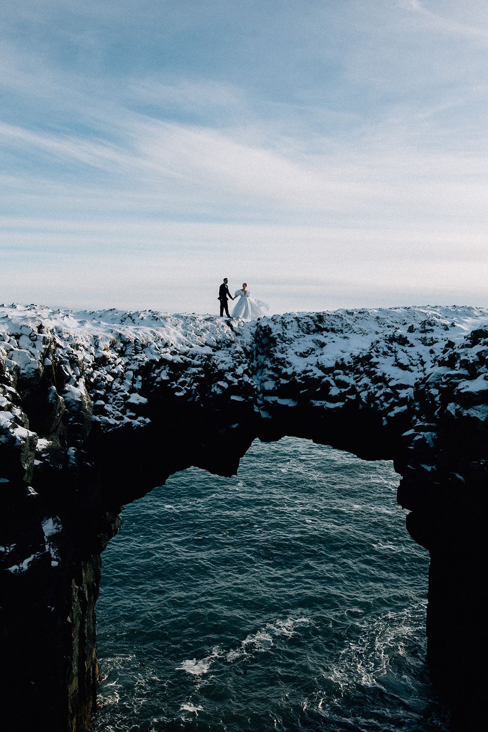 Bride and groom hold hands on snow-covered rocky arch over ocean in Iceland winter elopement photo.