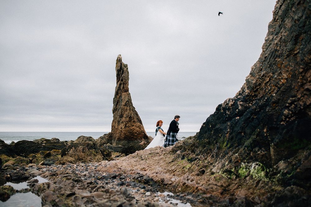 Couple in wedding attire walks along rocky Scottish coastline with dramatic sea stack and overcast skies.