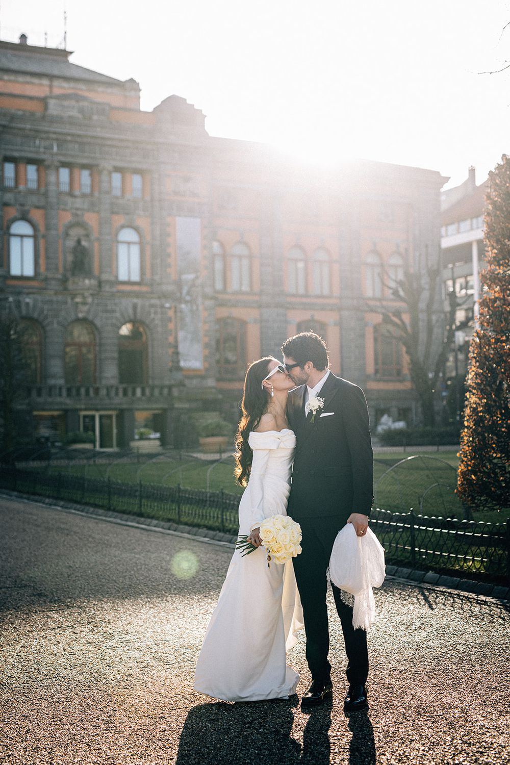 Bride and groom share a romantic kiss in golden sunlight outside a historic European building on their wedding day.