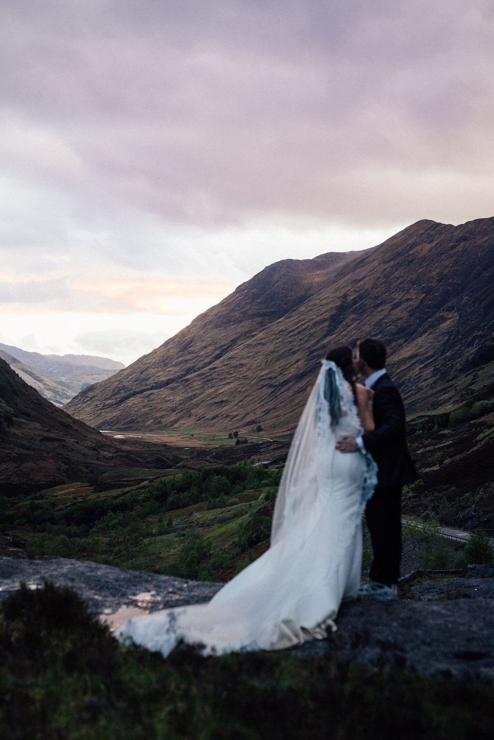 Bride and groom kissing at sunset in a Scottish Highland valley, with dramatic mountains and a flowing white veil.