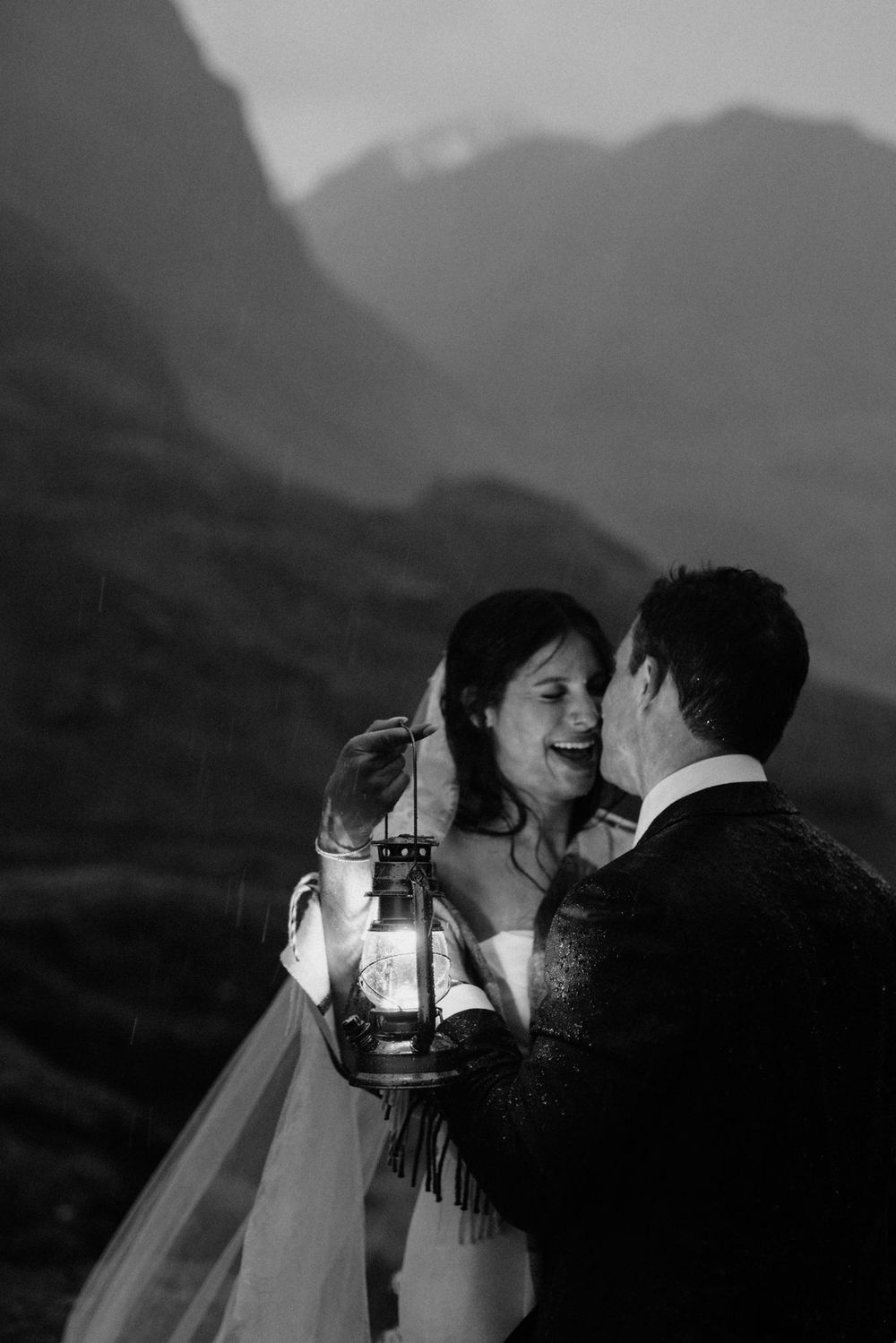 Black and white photo of a joyful bride and groom sharing a romantic moment while holding a glowing lantern with mountains behind.