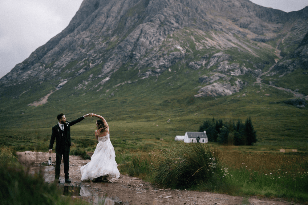 Bride and groom dancing in Scottish Highlands with dramatic mountain backdrop and white cottage.
