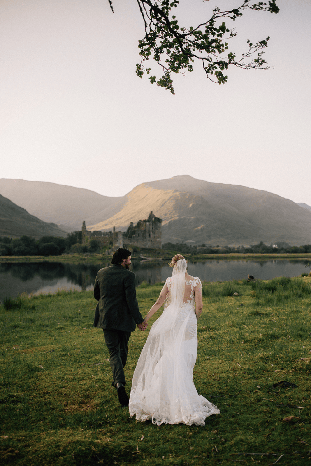 A bride and groom walk hand in hand toward a misty loch with ruins and mountains in the Scottish Highlands.