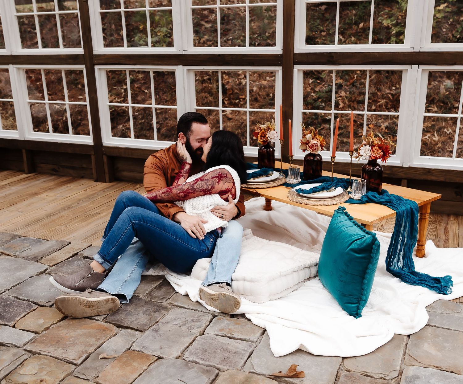Cozy indoor sunroom setup with blankets and pillows on stone floors next to large windowpanes.