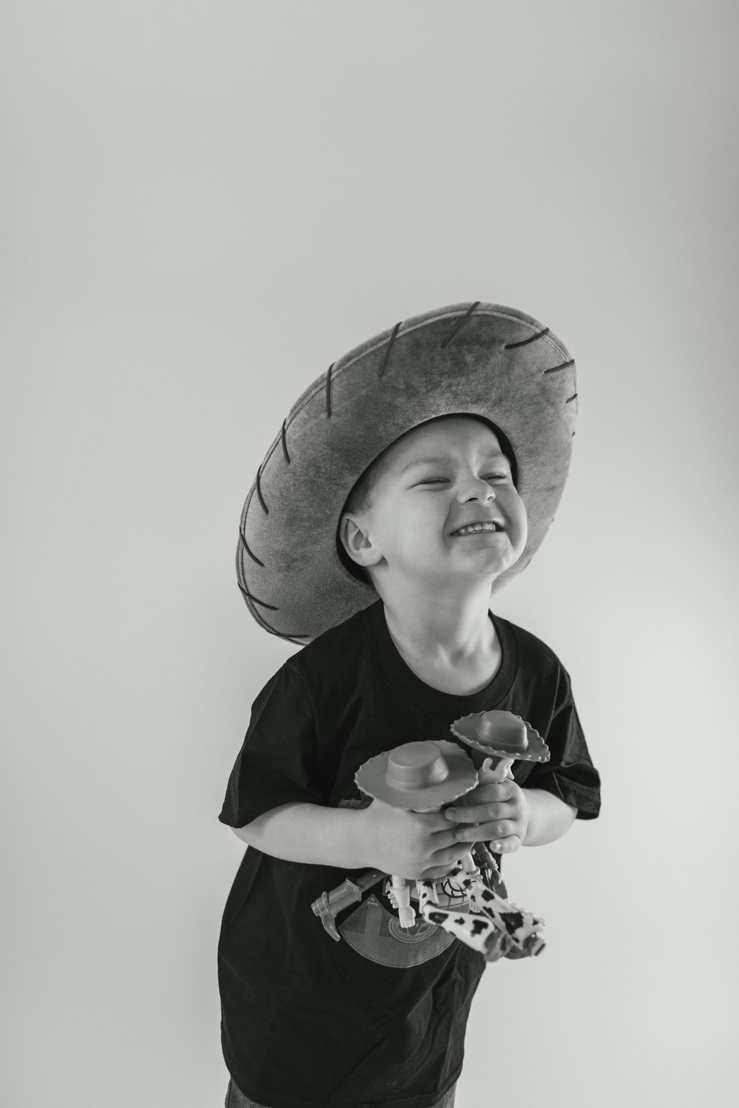 Child wearing a black t-shirt and cowboy hat laughs joyfully against a gray background.