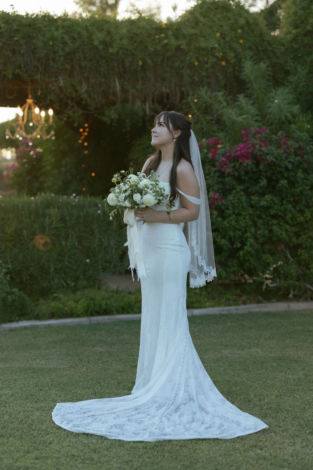 A bride in a fitted white lace gown with long veil holds a white bouquet in a garden setting at sunset.