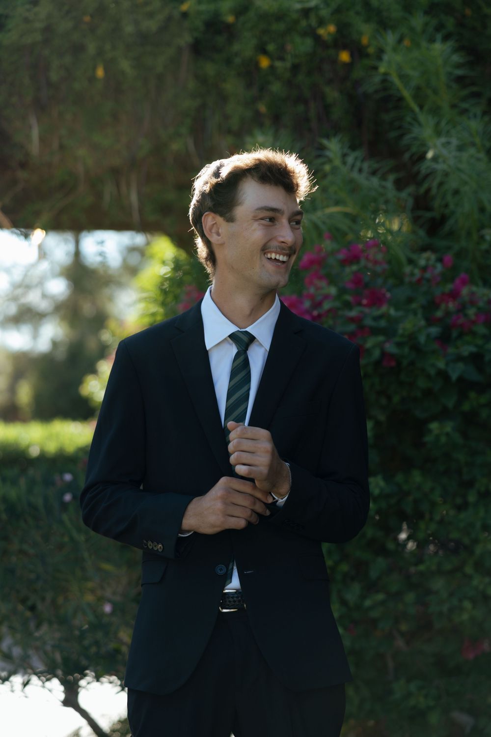 Groom in black suit laughing joyfully during outdoor wedding photos with greenery backdrop.