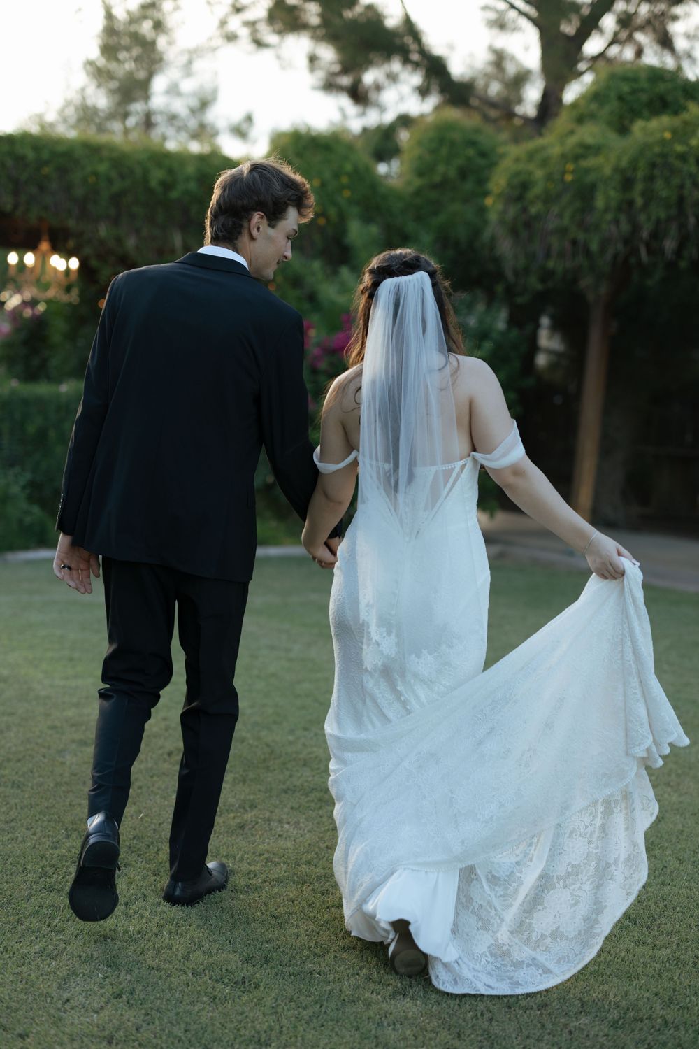 A couple walks together on grass, with the bride's white lace wedding dress trailing behind them in a romantic sequence.