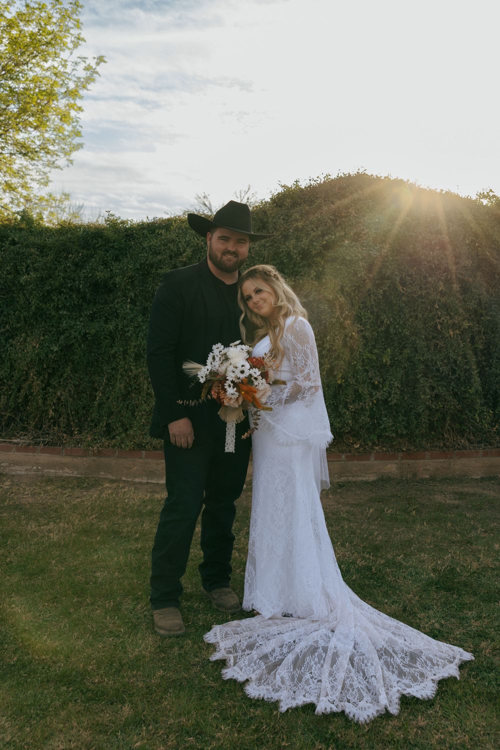 A bride in lace dress and groom in black stand together in a garden setting at sunset.