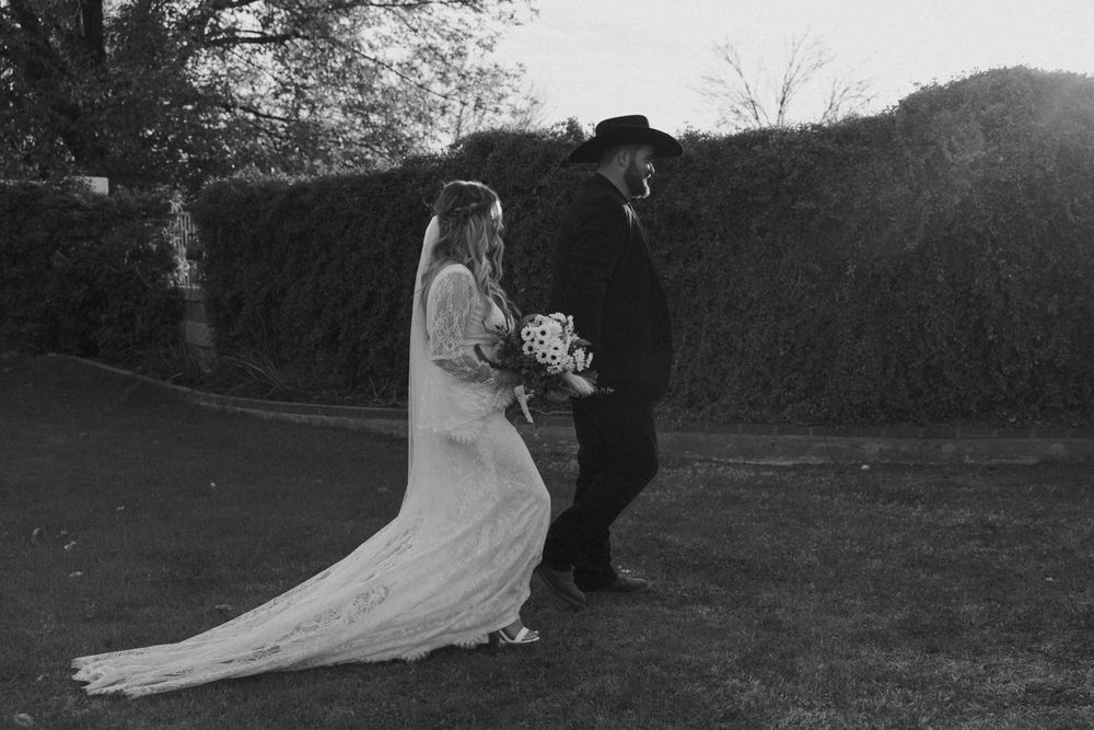 A black and white wedding photo of a couple in formal attire standing outdoors by a hedge.