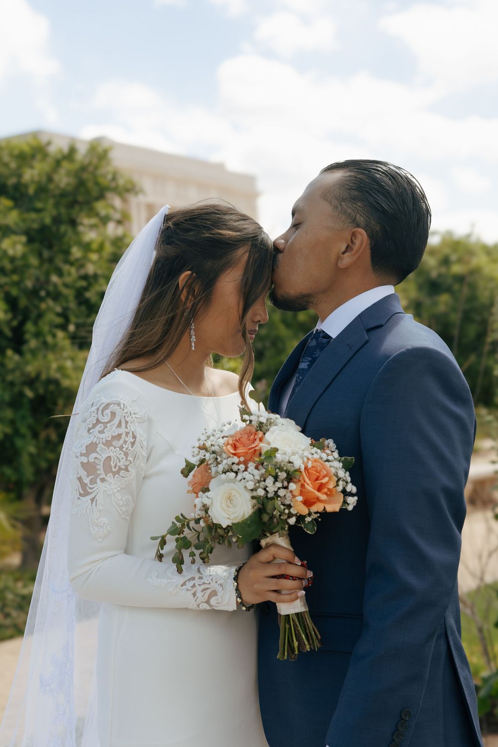 A romantic wedding couple embraces outdoors while the bride holds a beautiful peach and white floral bouquet.