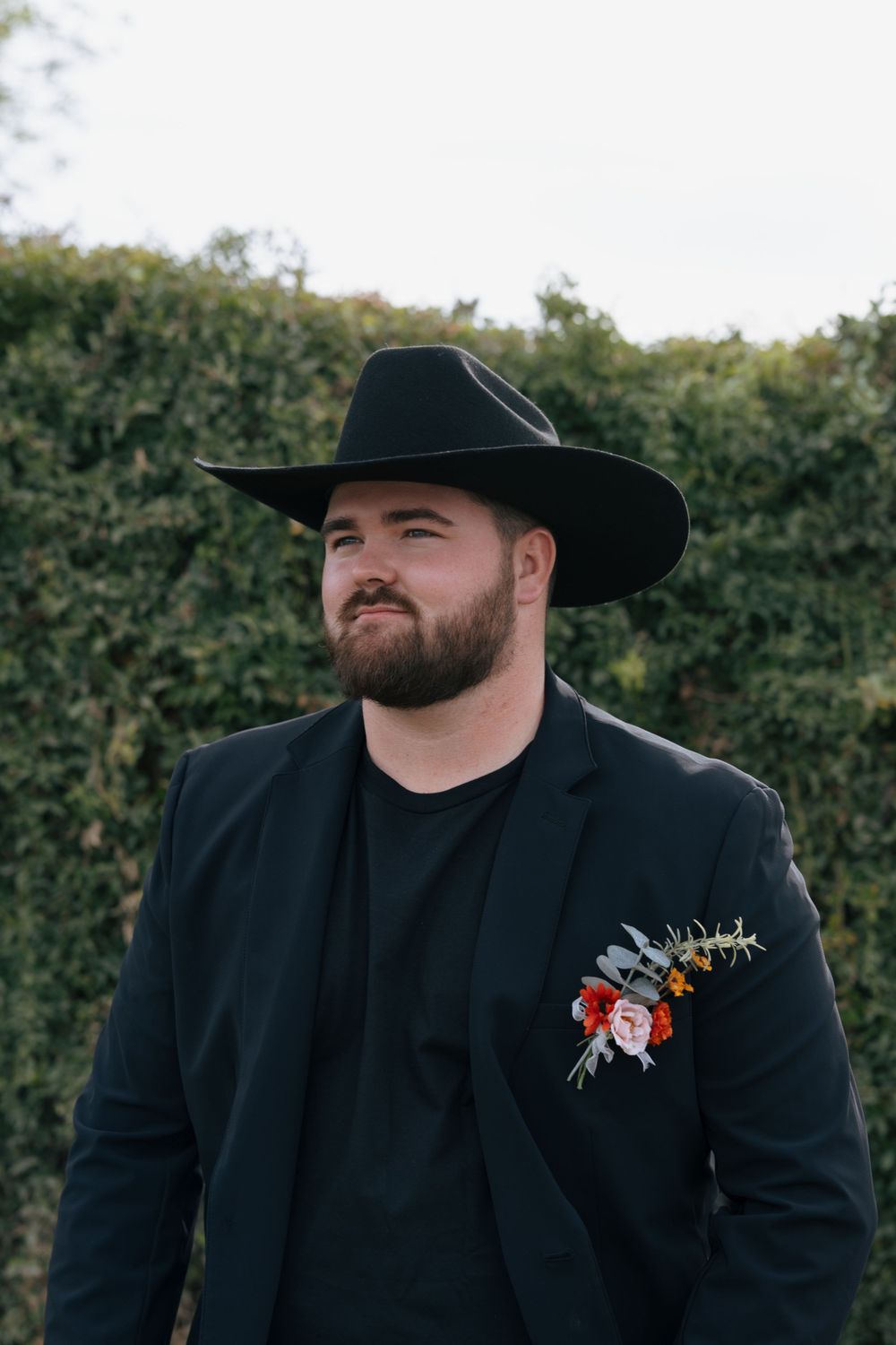 A groom in black western attire and cowboy hat smiles outdoors during a wedding.