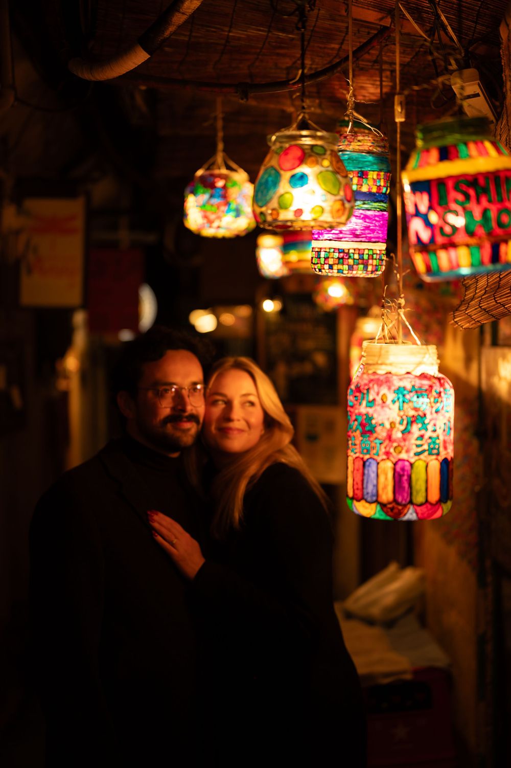 A couple embraces warmly beneath colorful hanging lanterns in a vibrant night market setting.