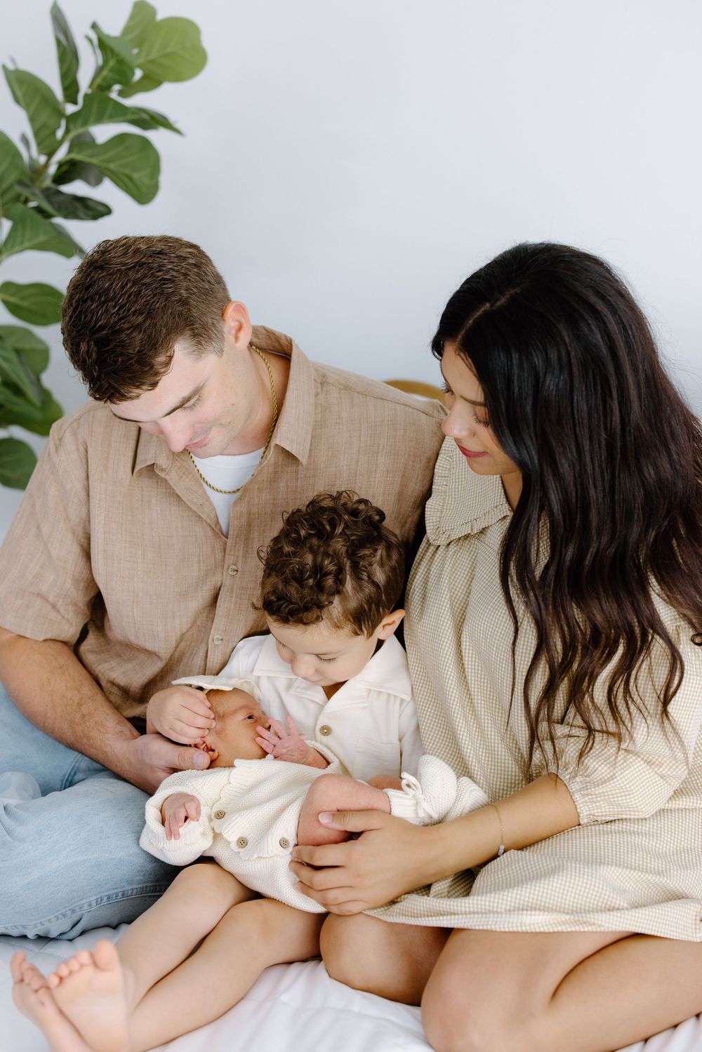 A young family sits together on a white bed, parents lovingly gazing at their newborn baby held by the mother.