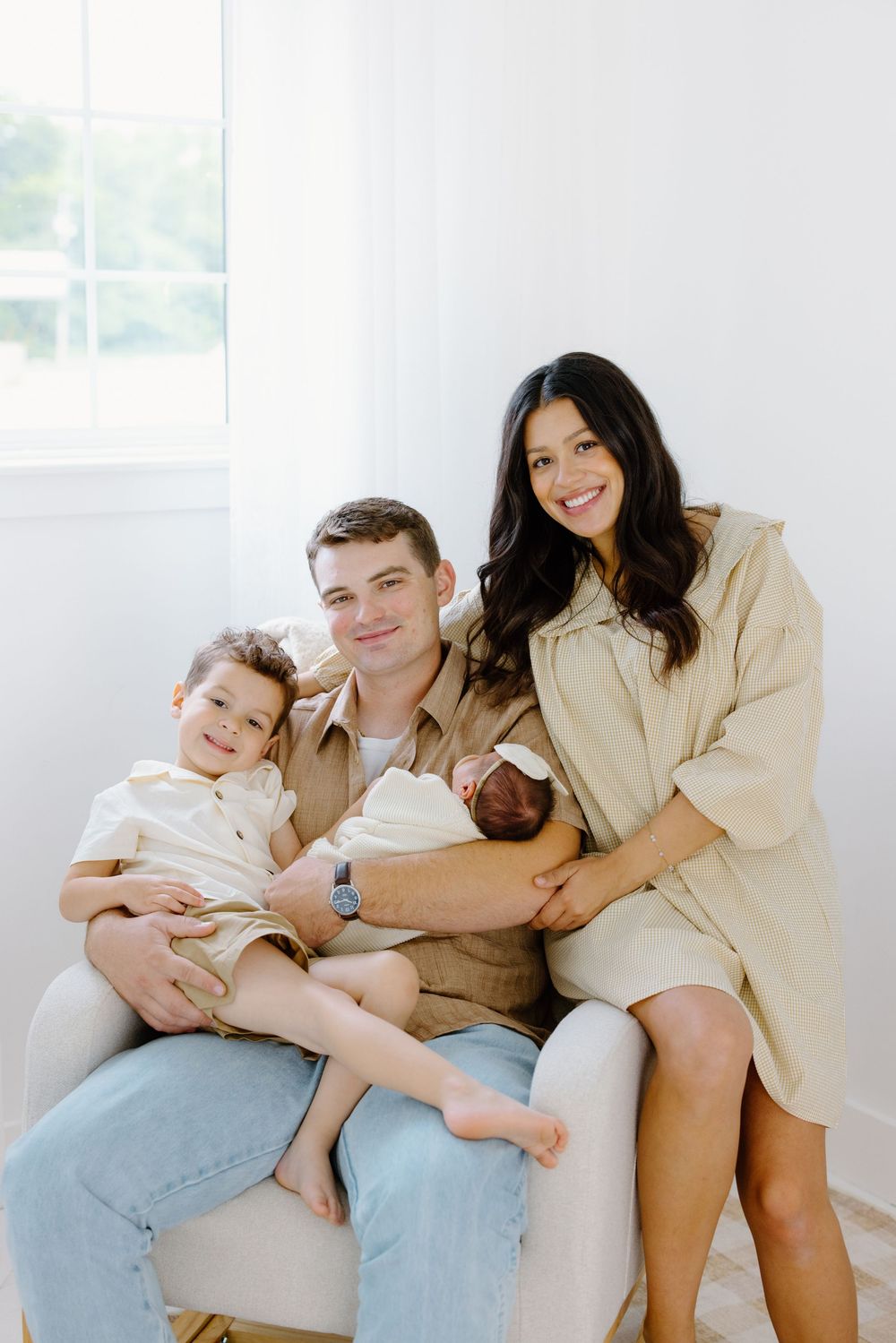 Happy family of four posing together, parents holding toddler and newborn baby in bright white room.