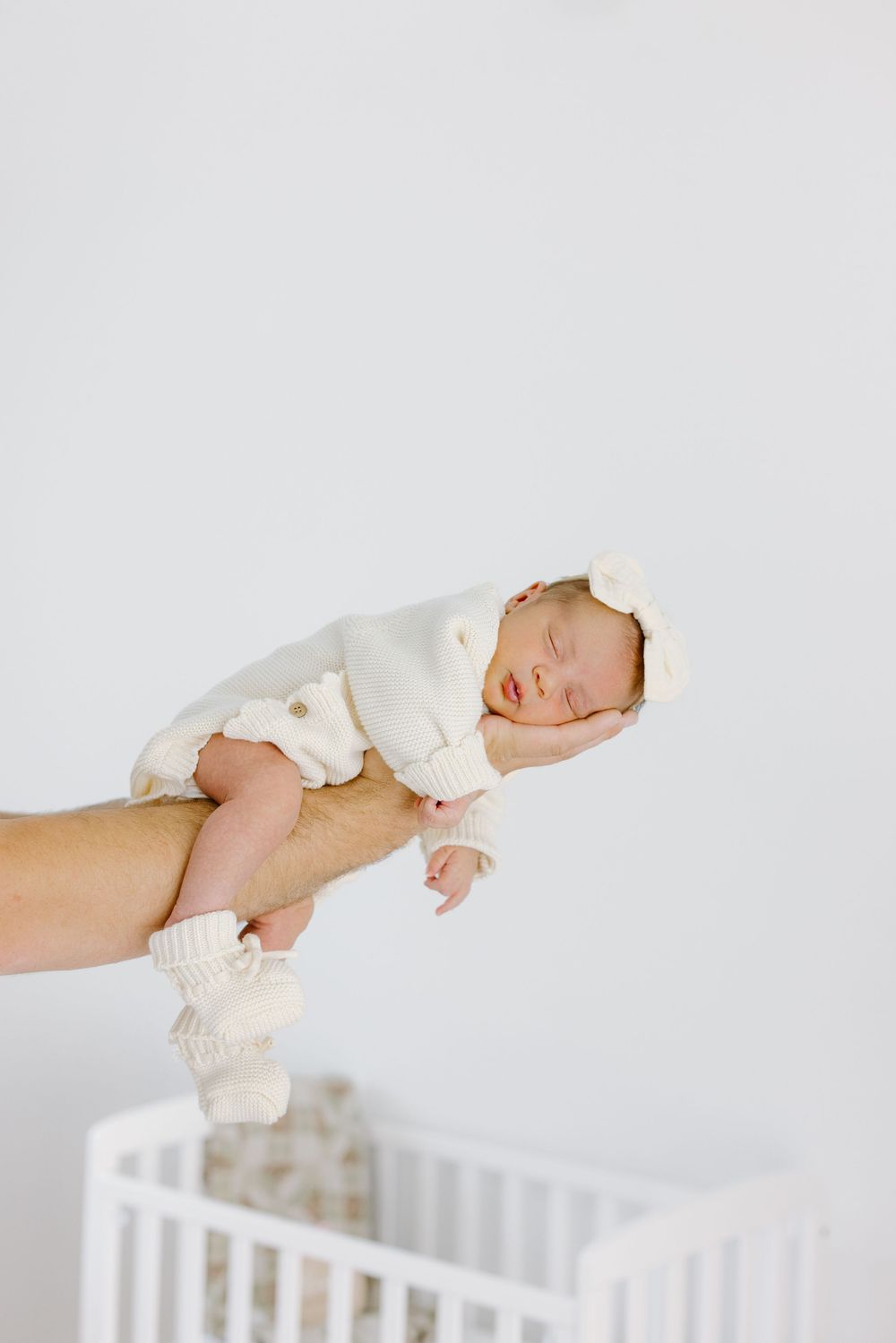 Newborn baby in white knit outfit held by adult hand over white crib, sleeping peacefully with bear ear headband.