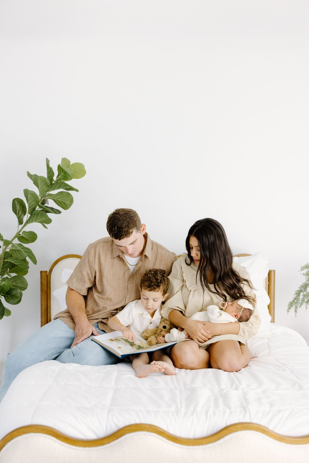 A family of three sits on a white bed, reading a book together, with a plant in the background.