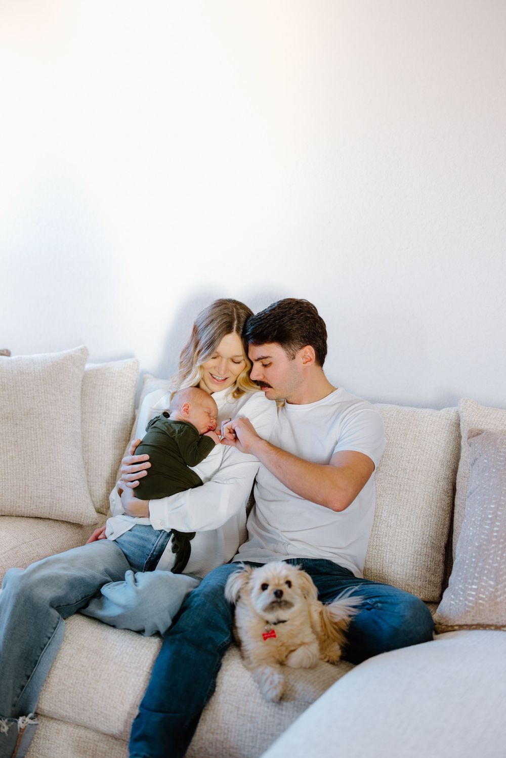 Young couple sitting on a beige sofa with their newborn baby and small white dog in a bright living room.