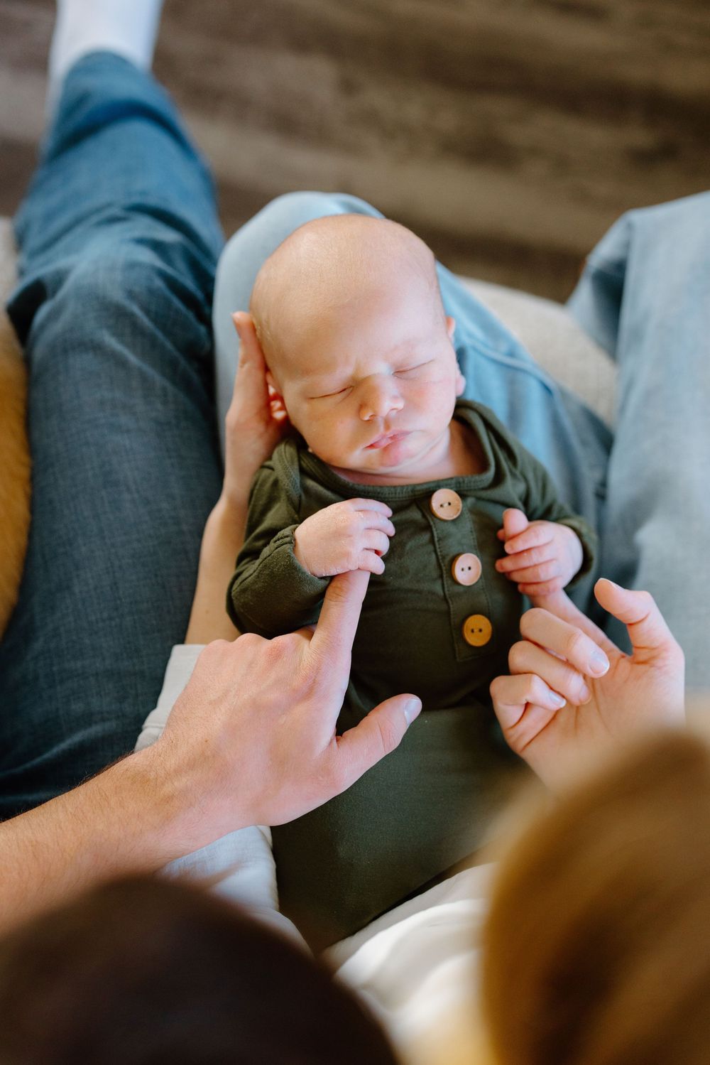 Sleeping newborn baby in green button onesie cradled in parent's arms while mother reaches out to touch.