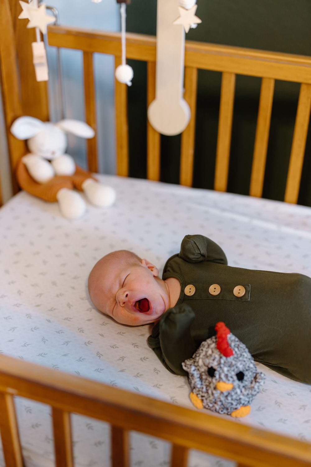 Newborn baby yawning in a wooden crib with a knit chicken toy and white bunny mobile above.