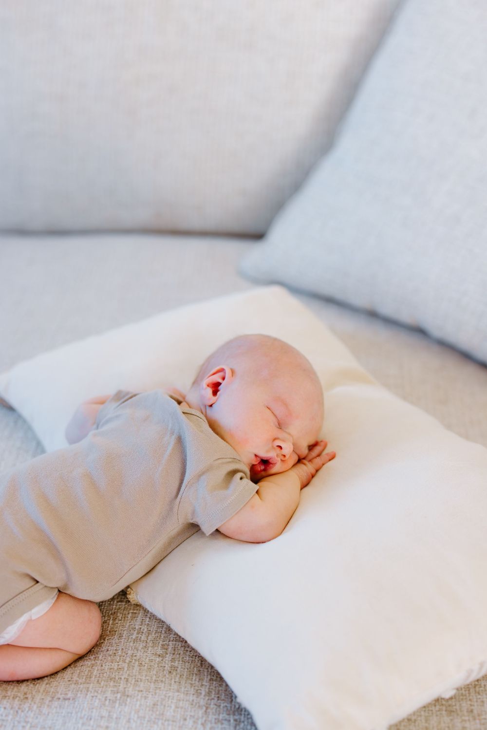 Peaceful newborn baby sleeping on a white pillow on a light gray sofa, wearing a soft gray onesie.