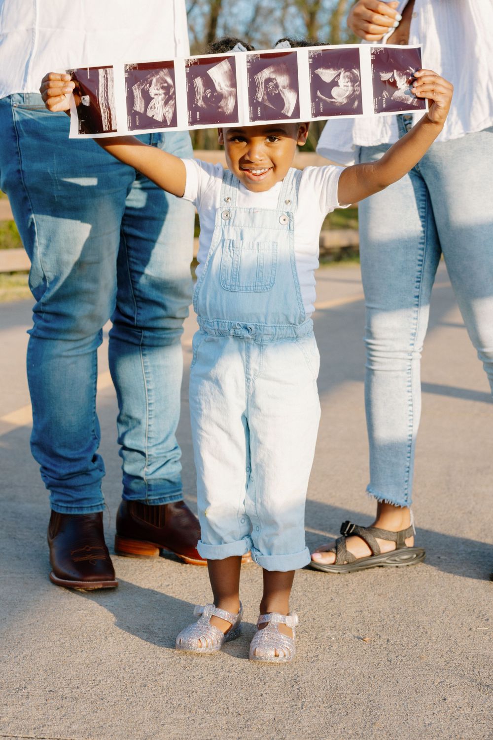 A child in white overalls holds a strip of ultrasound photos while standing between two people in jeans.
