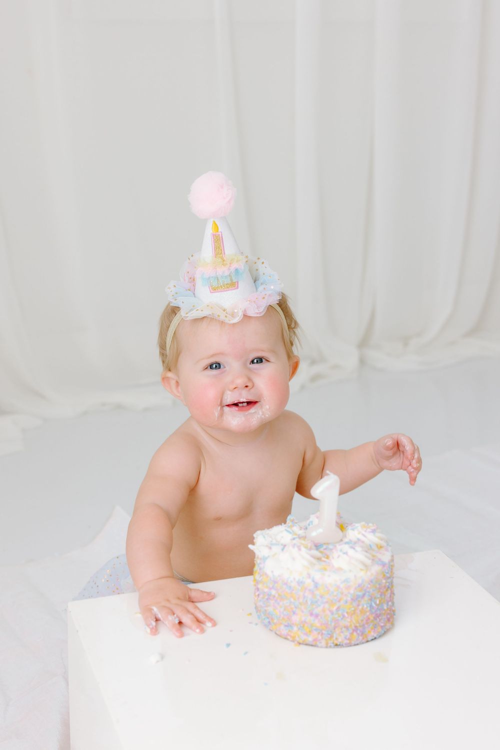 Smiling baby girl wearing a party hat celebrates her first birthday with a small white cake with one candle.