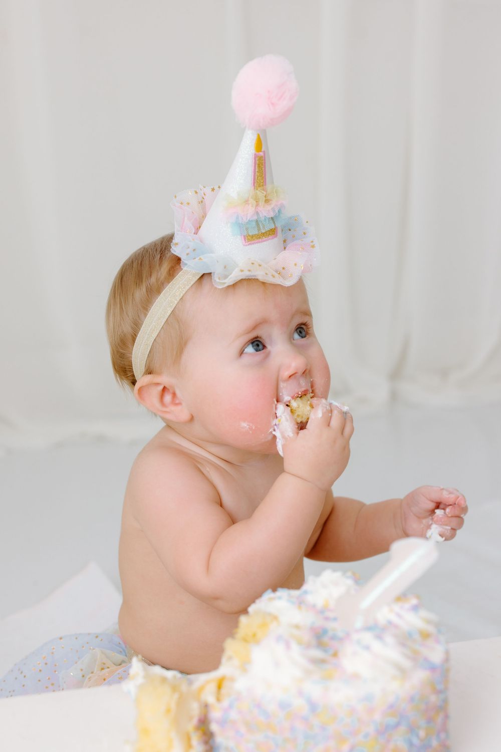Baby in party hat eating first birthday cake during a smash cake celebration on white background.