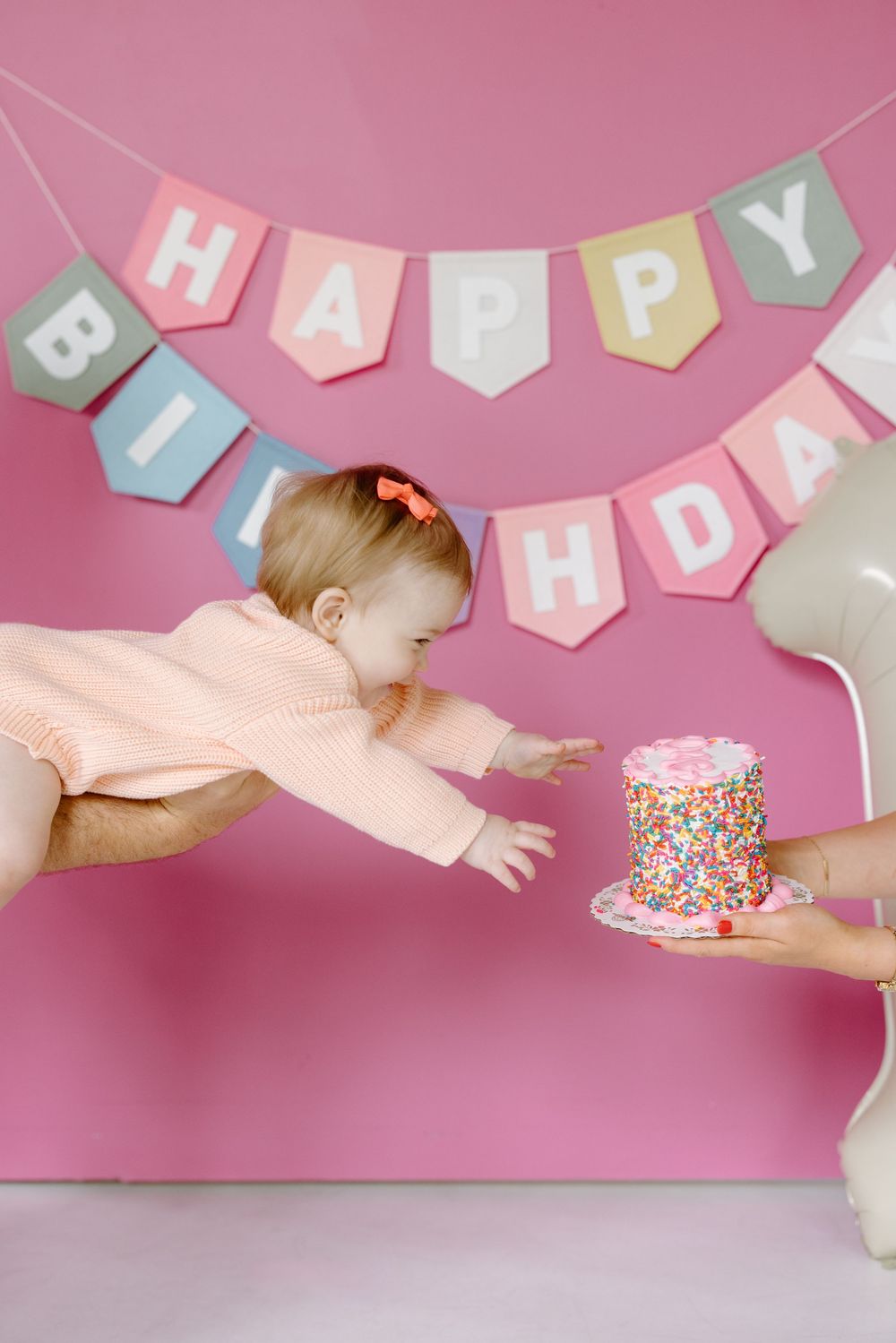 Baby reaching for colorful sprinkle birthday cake held by adult, floating in front of pink Happy Birthday banner backdrop.
