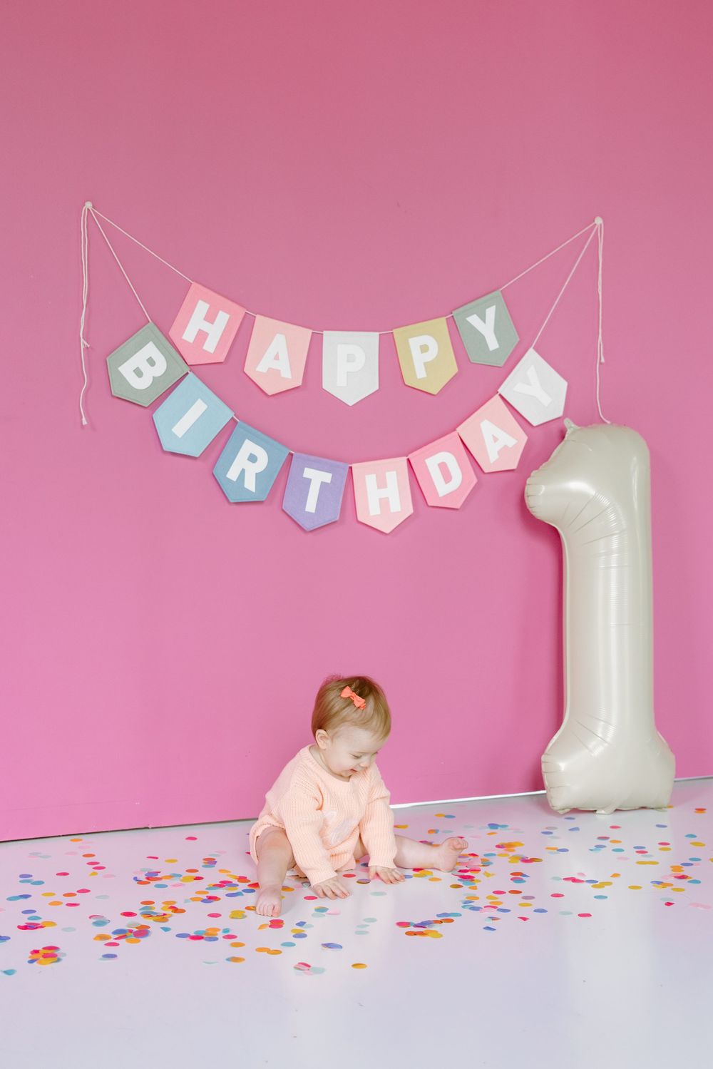Baby girl plays with confetti at first birthday party with Happy Birthday banner and number 1 balloon on pink wall.