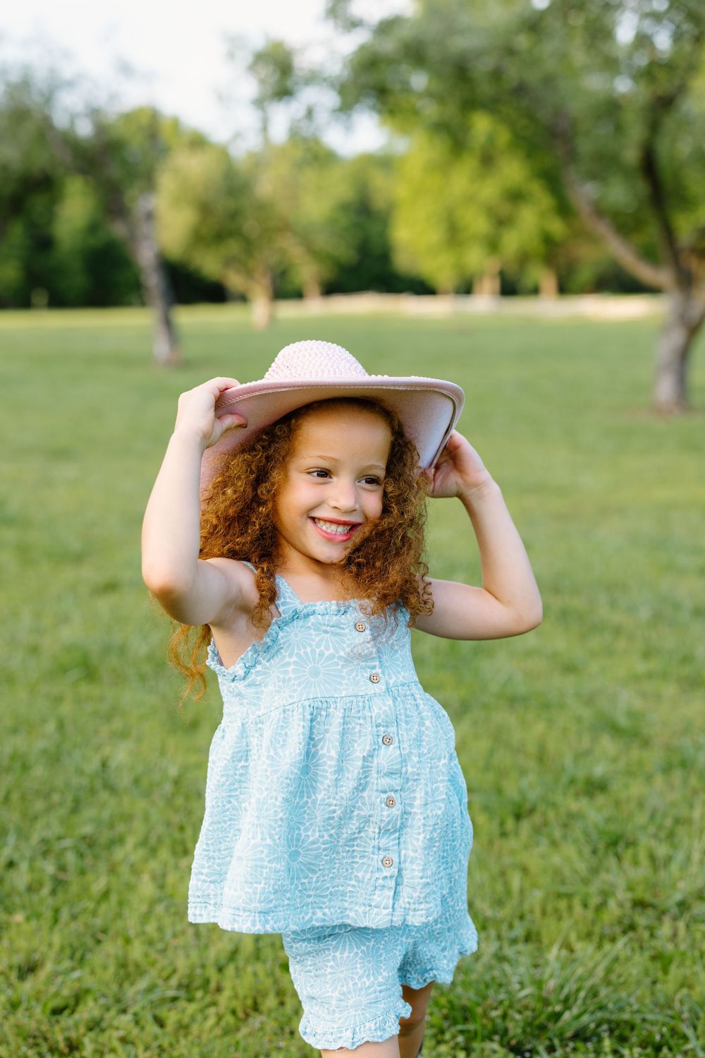Curly-haired girl in blue outfit holding pink sun hat, smiling in green park on sunny day.