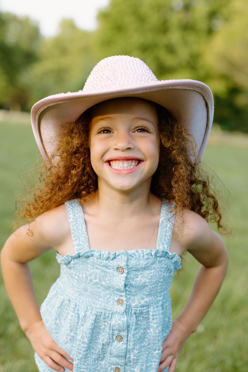 A cheerful young girl with curly red hair wearing a wide-brim hat and blue sundress smiling outdoors.