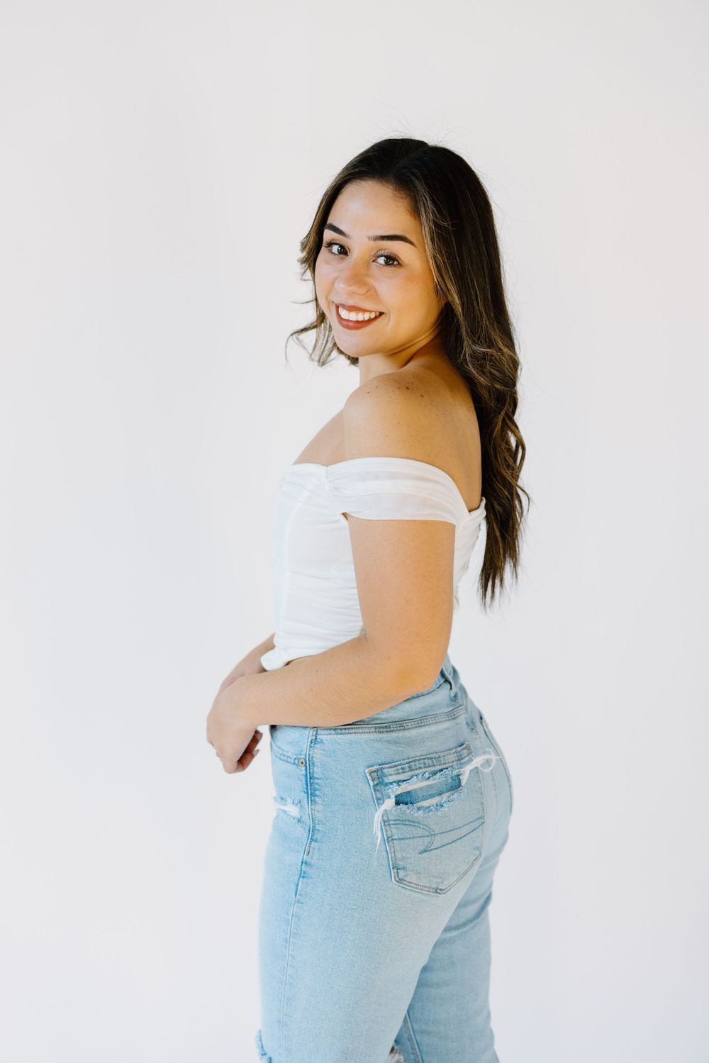A woman in a white off-shoulder top and light wash jeans poses against a white background.
