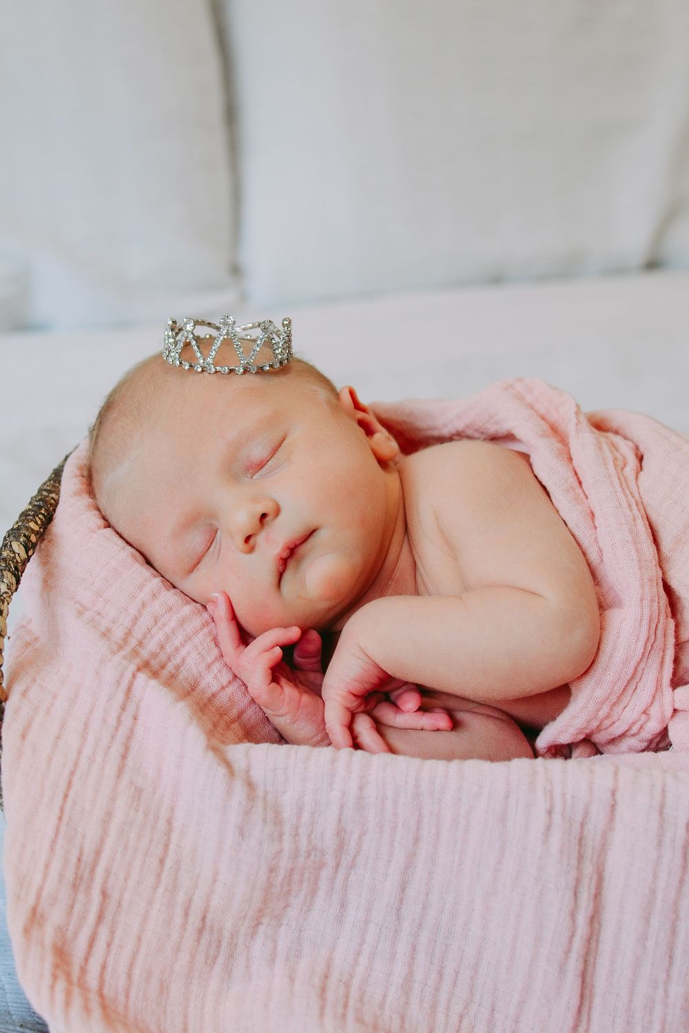 Sleeping newborn wearing delicate crown wrapped in soft pink muslin blanket on light background.