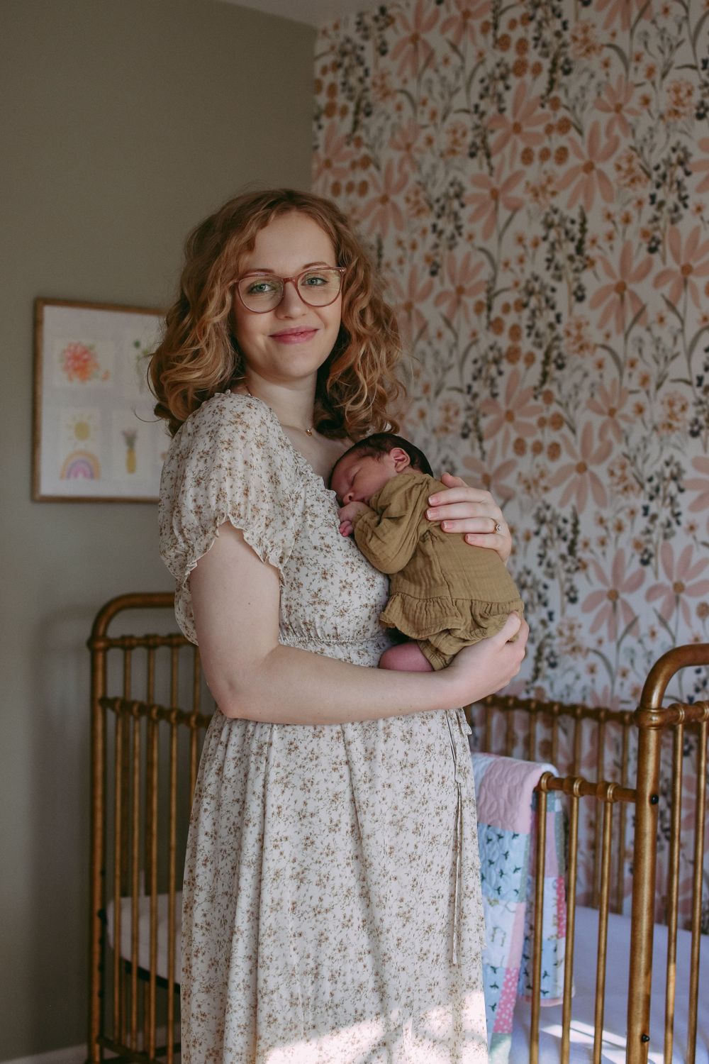 A person wearing glasses and a floral dress holds a newborn baby in a nursery with floral wallpaper and wooden crib.