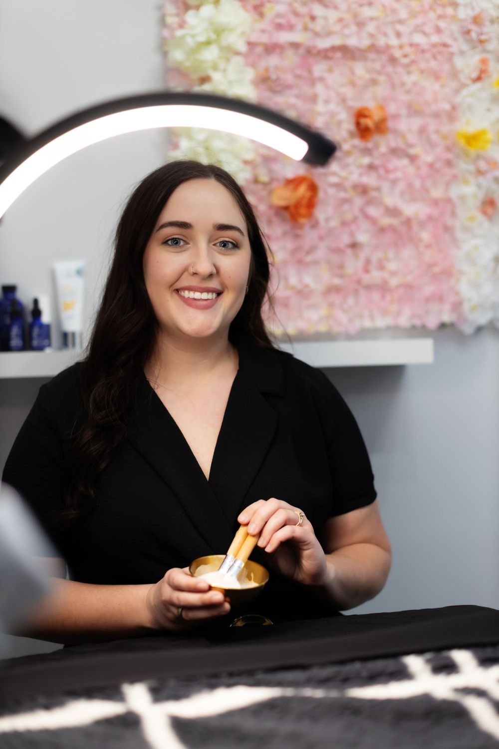 Professional wearing a black wrap top smiles while working at a counter with floral decor in the background.