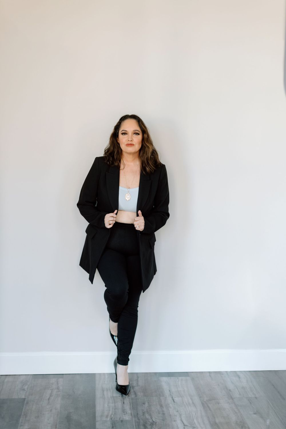 A person in a professional black suit and pants ensemble poses against a white wall with gray wood flooring.