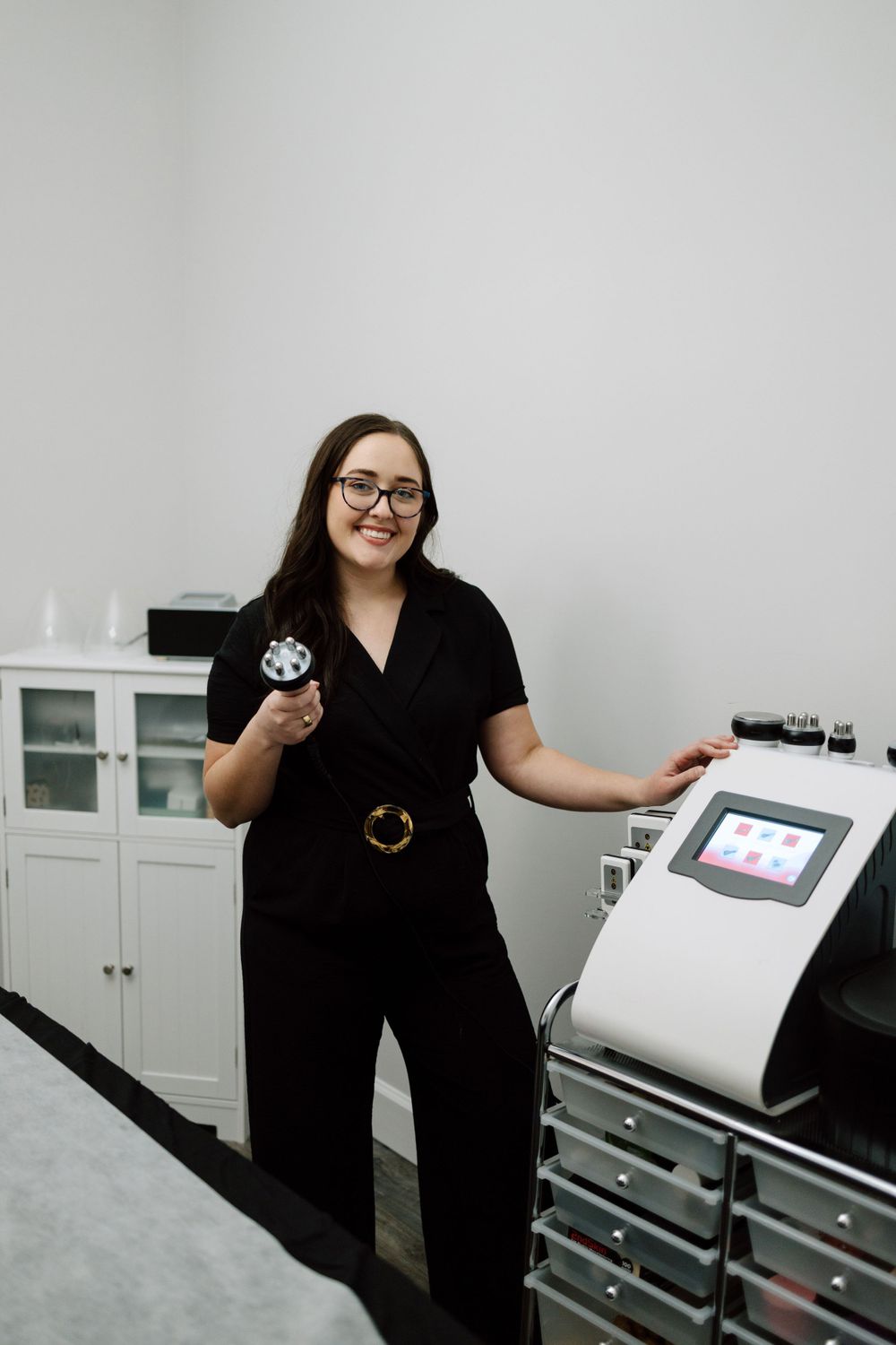Healthcare professional in black scrubs stands next to medical equipment in a clinical setting.