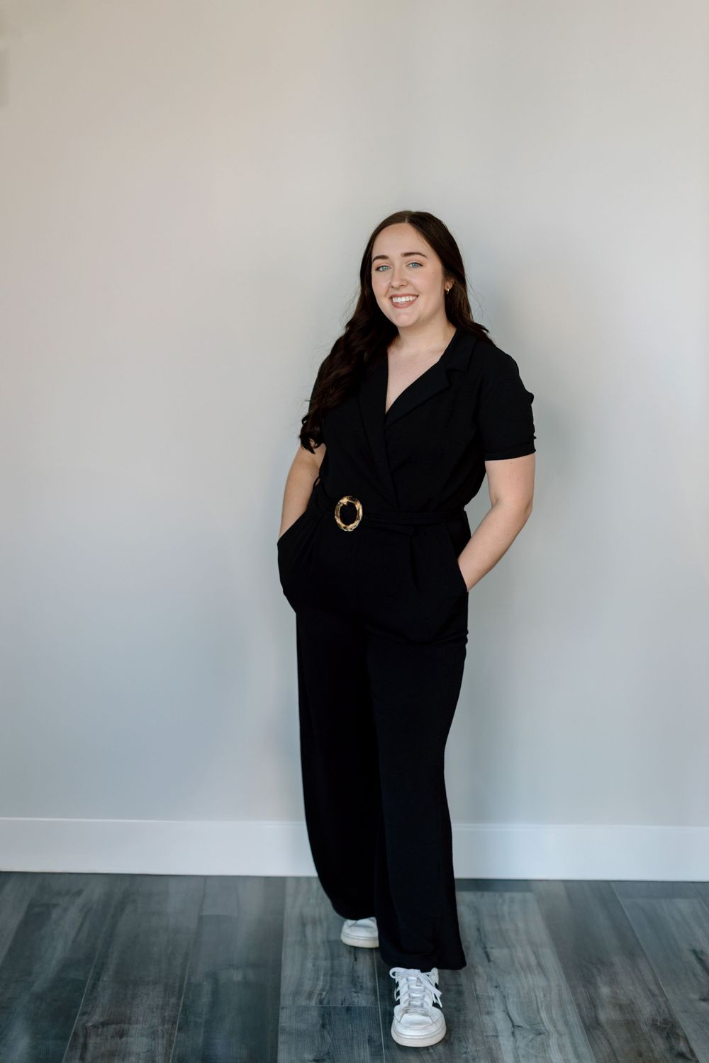 Person in a black jumpsuit with belt detail poses casually against a gray wall while wearing white sneakers.
