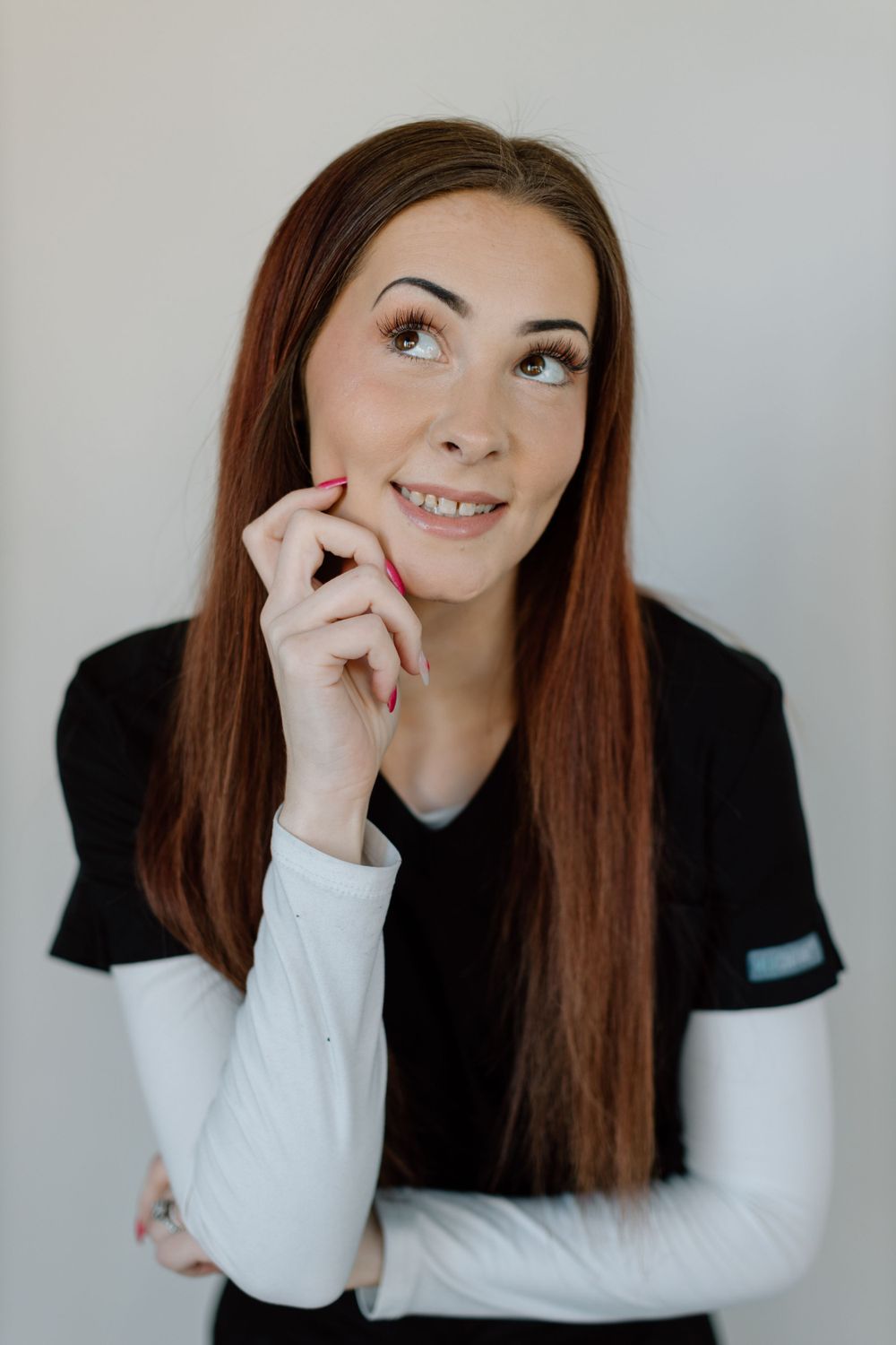 A thoughtful young person with long brown hair wearing a black and white top looks upward while touching their face.
