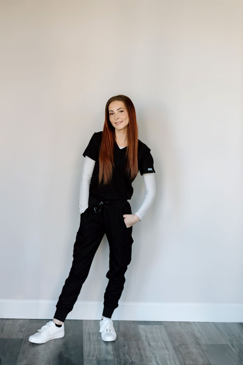 Someone in an all-black casual outfit with long brown hair poses against a white wall in a minimalist interior.
