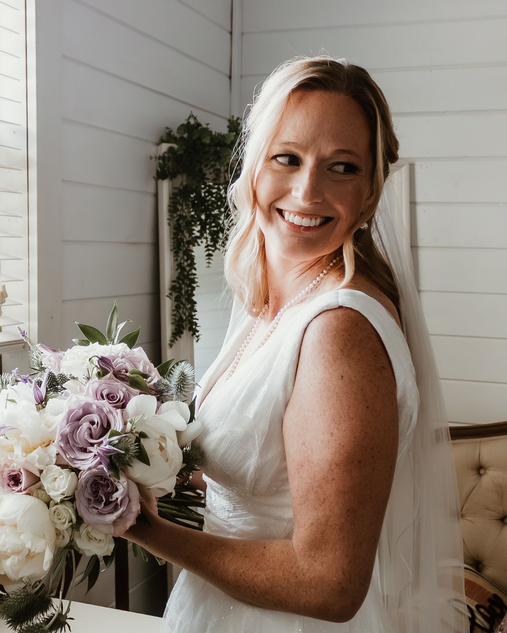 A bride in a classic white gown holds a beautiful purple and white bouquet while smiling beside a white wall.