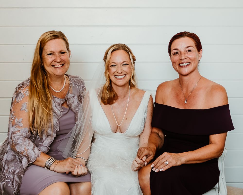 Three women pose together at a wedding, with the bride in white between two guests in formal attire.