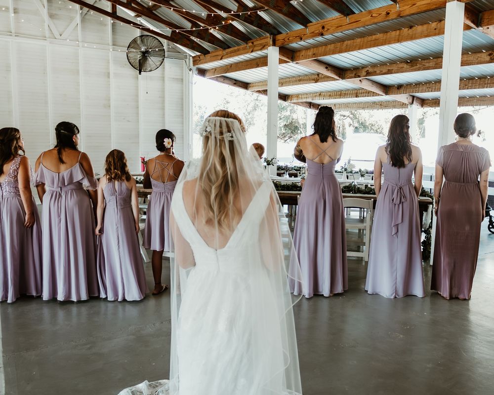 Bridesmaids in lavender dresses stand in a row while the bride in white faces them from behind.